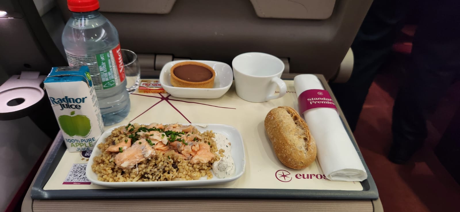A meal on board Eurostar Standard Premier. There is a water bottle, apple juice, a salmon salad, a bread roll, a chocolate tart and a hot drink.